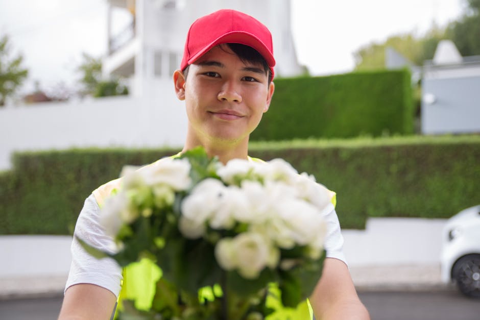 A young male flower delivery courier wearing a red cap and a high-visibility vest holds a bouquet of fresh white roses and green foliage, extending it towards the camera with a friendly smile. He stan