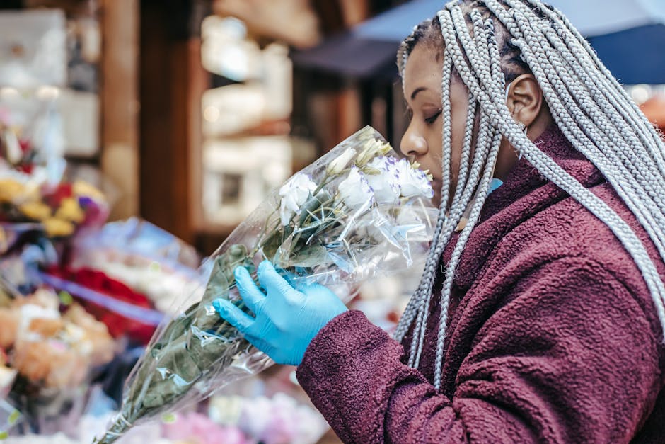 A person with silver and purple braided hair wearing a maroon fleece jacket and blue disposable gloves is holding a bouquet of fresh white roses wrapped in clear plastic. They are at an outdoor flower