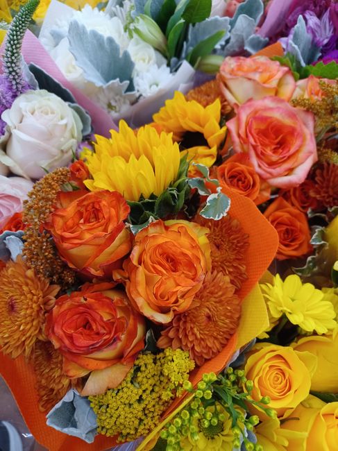 A vibrant floral bouquet arranged with a variety of fresh flowers, including orange and peach roses, bright yellow chrysanthemums, soft pink roses, and purple lavender accents. The flowers are wrapped