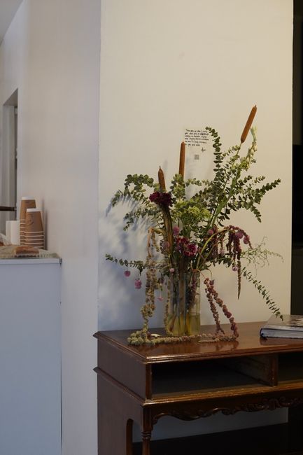 A tall, artistic floral arrangement in a clear glass vase, featuring various flowers and greenery, including prominent pink and purple blossoms, accented with hanging and trailing foliage. The arrange