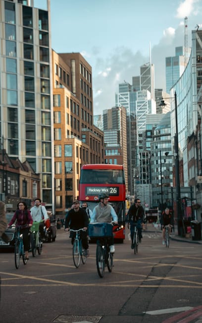 A busy street scene in Hackney Wick featuring pedestrians walking and cycling transportation against a backdrop of modern high-rise buildings with glass facades reflecting the sky. A red double-decker