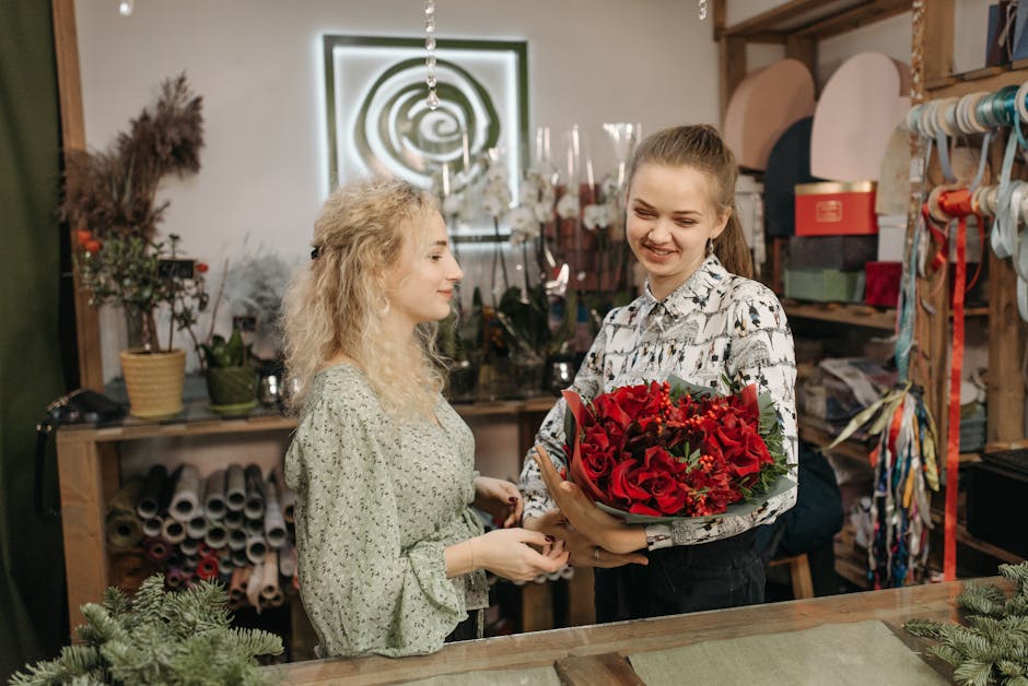 A young woman with light brown hair tied back, smiling as she hands over a vibrant bouquet of red roses, lilies, and greenery wrapped in green floral paper, to another woman with curly blonde hair wea